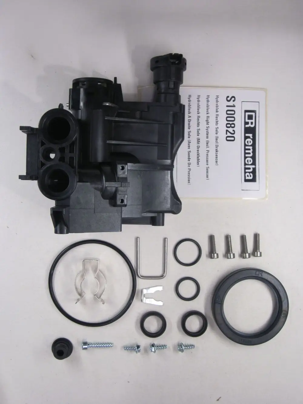 Hydraulikblock der Marke Remeha mit Anschluss für Druckfühler, umgeben von verschiedenen Dichtungsringen, Schrauben und Befestigungselementen auf weißem Hintergrund.